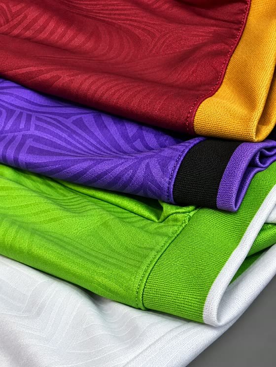Quatro uniformes dobrados mostrando o relevo tridimensional do tecido em vinho, roxo, verde e branco
